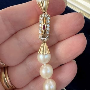 May include: A pearl necklace with a gold clasp featuring a central ruby and surrounding clear gemstones. The necklace has multiple creamy white pearls. The clasp has a rectangular shape with a gold frame.