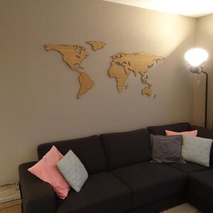 Può includere: Una mappa del mondo in legno è appesa a una parete neutra sopra un divano angolare grigio scuro. Il divano è decorato con diversi cuscini rosa, grigi e verde chiaro. Una lampada da terra con una grande luce rotonda è nell'angolo.
