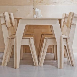 Pode incluir: Um conjunto de jantar em madeira clara com uma mesa e quatro cadeiras. As cadeiras têm um design único com pernas e encostos angulados. A mesa é retangular e o conjunto está numa sala com paredes claras.