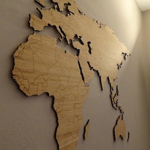 Peut inclure: Une grande carte du monde en bois, fixée sur un mur gris texturé. La carte est en bois clair, avec des contours détaillés des continents et des pays. La carte est éclairée par une lampe.