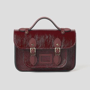 Può includere: Una borsa a cartella bordeaux lucida, con manico superiore e patta frontale fissata da due fibbie. La borsa presenta una piccola tasca frontale ed è realizzata in un materiale lucido e strutturato. L'hardware è color oro.