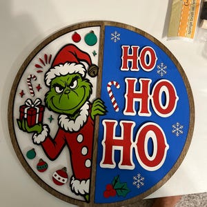 Könnte beinhalten: Ein rundes Holzschild, das in zwei Hälften unterteilt ist. Die linke Seite zeigt eine grüne Figur im roten Weihnachtsmannkostüm mit einem Geschenk, umgeben von kleinen Ornamenten und einer Zuckerstange. Die rechte Seite ist blau mit rotem Text HO HO HO.