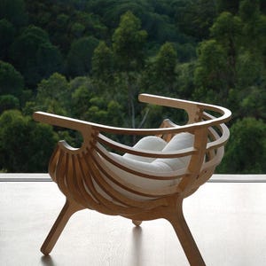 Könnte beinhalten: Ein moderner Holzstuhl mit einzigartigem Design. Der Stuhl hat eine geschwungene Rückenlehne und Armlehnen, wobei vertikale Holzlatten die Rückenlehne des Sitzes bilden. Ein weißes Kissen bietet Komfort. Der Stuhl ist aus hellem Holz gefertigt.