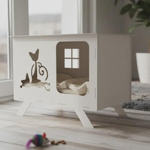 Peut inclure: Une maison pour chat blanche avec une silhouette de chat découpée et une fenêtre. La maison a une forme rectangulaire avec des pieds inclinés et un intérieur douillet avec une couverture. Une souris jouet est au sol.