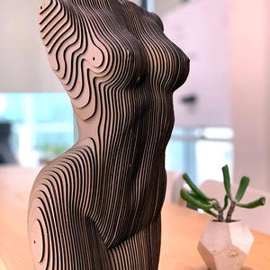 May include: A wooden sculpture of a female torso, constructed from layered horizontal lines in alternating light and dark brown. The sculpture has a striped appearance, with a smooth, contoured form. A small succulent plant in a geometric pot is placed nearby. The sculpture is approximately 30 cm tall.
