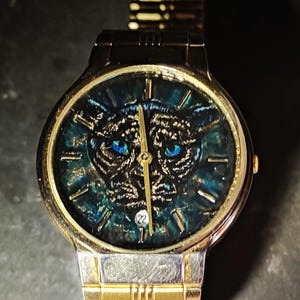 Puede incluir: Un reloj de pulsera analógico de tono dorado con correa de eslabones metálicos. La esfera presenta un fondo texturizado en azul verdoso oscuro y negro con una ilustración central de una cabeza de pantera, ojos azul brillante, manecillas doradas y una ventana de fecha.