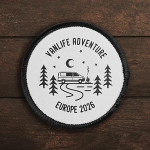 May include: Circular patch with a black border and a white centre. It features a van, trees, a campfire, and the text "VANLIFE ADVENTURE EUROPE 2026". The design is in black and white, set against a dark wood grain background.