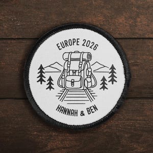 May include: A circular white embroidered patch with a black border featuring a black line art illustration of a backpack on train tracks, pine trees, and mountains. The text reads EUROPE 2026 at the top and HANNAH & BEN at the bottom.