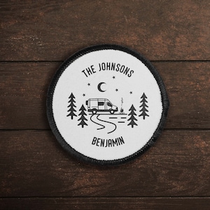 May include: A white round patch with black stitching. The patch features a black and white illustration of a camper van driving through a forest with a campfire and a crescent moon in the sky. The text "THE JOHNSONS" is above the illustration and "BENJAMIN" is below.