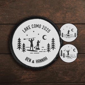 Könnte beinhalten: Ein weißer runder Patch mit schwarzem Text und Design. Der Text lautet "LAKE COMO 2025" mit zwei Sternen und einem Halbmond über dem Text. Unter dem Text befindet sich eine Silhouette von zwei Personen beim Paddelsurfen mit einem Halbmond im Hintergrund. Der Text "BEN & HANNAH" befindet sich unter den Paddelsurfern. Der Patch hat einen schwarzen Rand.