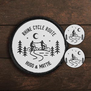 Pode incluir: Um remendo branco redondo com texto preto e uma silhueta preta de duas pessoas andando de bicicleta em um caminho sinuoso sob uma lua crescente. O texto diz "Rhine Cycle Route Hugo & Mattie".