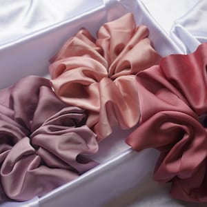 May include: Three satin scrunchies in shades of pink, mauve, and burgundy. The scrunchies are arranged in a white box.