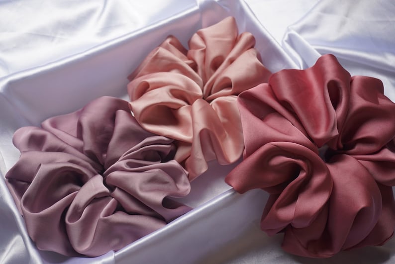 May include: Three satin scrunchies in shades of pink, mauve, and burgundy. The scrunchies are arranged in a white box.
