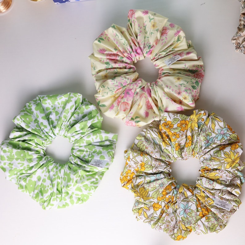May include: Three fabric scrunchies with floral patterns. The scrunchies are in shades of green, yellow, and pink. Each scrunchie has a small white tag with the text "CREA A FUNK" on it.