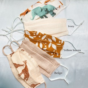 May include: Five fabric face masks in various designs. One mask features animal illustrations, another is ivory, a third has yellow leaves, a fourth is beige, and the last one has giraffe patterns. Each mask has adjustable white ear loops.