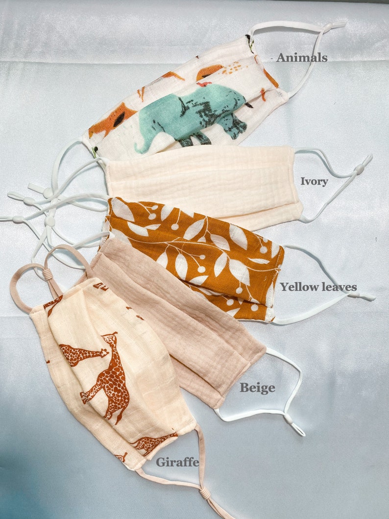 May include: Five fabric face masks in various designs. One mask features animal illustrations, another is ivory, a third has yellow leaves, a fourth is beige, and the last one has giraffe patterns. Each mask has adjustable white ear loops.
