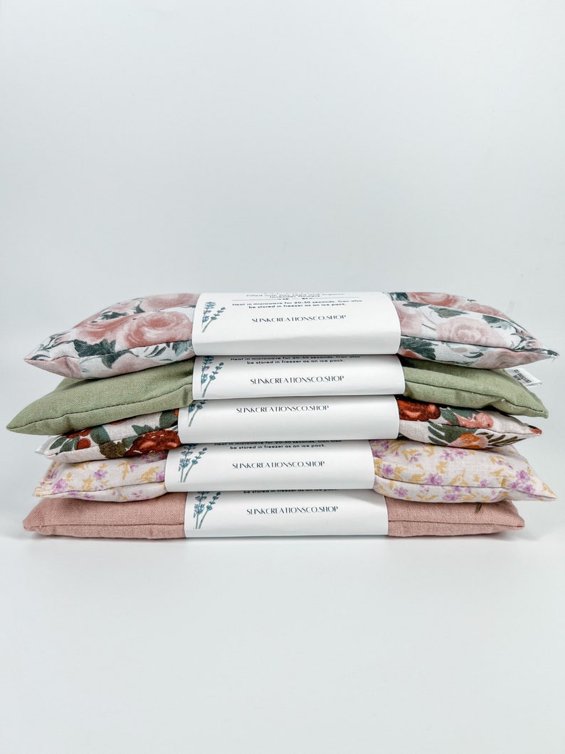 May include: A stack of five floral eye pillows, each with a different pattern. The pillows are all pink, green, and white. The pillows are labelled "SunKissed Aromas Co. Shop".