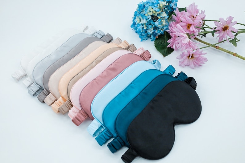 May include: A set of ten silk sleep masks in various colours, including white, grey, beige, pink, blue, teal, and black. The masks are arranged in a fan shape, with the black mask at the bottom.