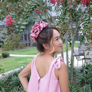 May include: A woman wearing a pink sleeveless dress with a bow detail on the straps. She has a large pink scrunchie in her hair.
