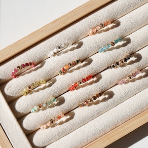 Puede incluir: Un expositor de madera con 10 anillos coloridos hechos de pequeñas piedras. Los anillos están dispuestos en filas sobre un fondo de tela beige.