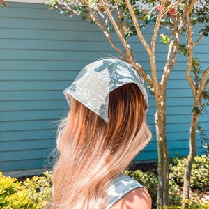 Op de afbeelding: Een vrouw met lang blond haar die een groene en witte bandana met bloemenprint om haar hoofd heeft gebonden.