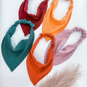 May include: Five fabric headbands in different colors: burgundy, orange, green, rust, and pink. The headbands are made of a soft, textured fabric and are tied at the back of the head.