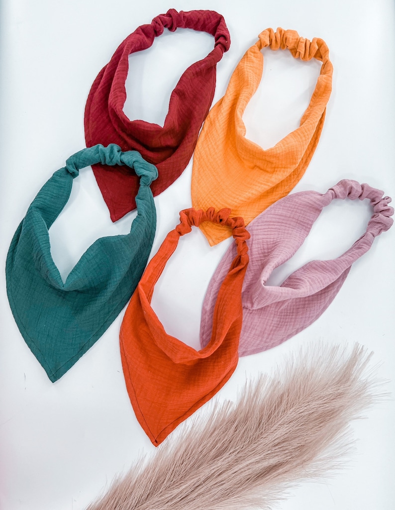 May include: Five fabric headbands in different colors: burgundy, orange, green, rust, and pink. The headbands are made of a soft, textured fabric and are tied at the back of the head.
