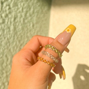 May include: Three gold, silver, and green beaded rings stacked on a finger. The rings are made of wire and have a braided design.