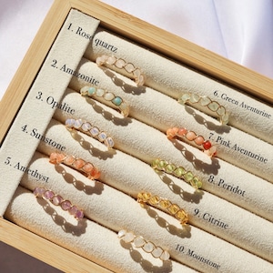 May include: Ten gemstone rings displayed in a wooden box. The rings are made of gold wire and feature different coloured gemstones, including rose quartz, amazonite, opalite, sunstone, amethyst, green aventurine, pink aventurine, peridot, citrine, and moonstone.