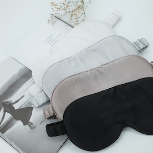 May include: Four silk sleep masks in different colours: white, light grey, beige, and black. The masks are arranged in a stack, with the black mask on top.