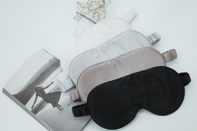 May include: Four silk sleep masks in different colours: white, light grey, beige, and black. The masks are arranged in a stack, with the black mask on top.