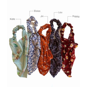 May include: Five colorful hair scrunchies with floral patterns. The scrunchies are arranged in a row from left to right: green with white flowers, blue with white flowers, orange with white flowers, purple with white flowers, and red with white flowers.