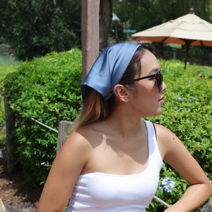 May include: A woman wearing a light blue bandana, sunglasses, and a white one-shoulder top. She is standing in front of a green bush with blue flowers.