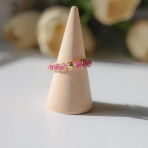 May include: A gold-colored wire ring with pink beads and a gold bead in the center. The ring is displayed on a light brown cone-shaped ring holder. The background is blurred, with hints of yellow flowers.