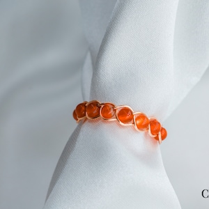 May include: A carnelian gemstone ring with a wire wrapped design. The ring is made with orange carnelian beads and is set on a white background.