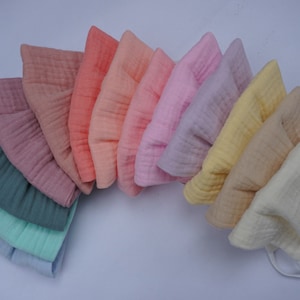 May include: A collection of ten muslin bonnets in a variety of pastel colors, including pink, yellow, and blue. Each bonnet has a soft, textured appearance and is arranged in a fan shape. The bonnets are likely for babies or young children.