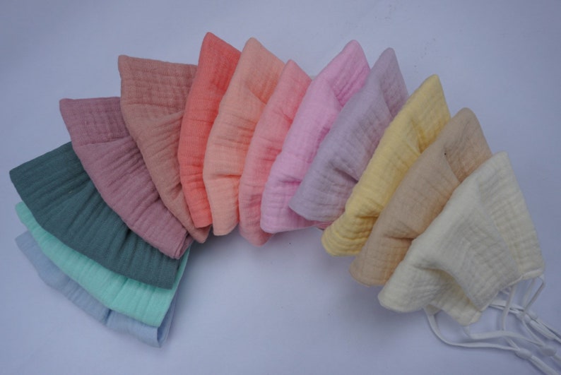 May include: A collection of ten muslin bonnets in a variety of pastel colors, including pink, yellow, and blue. Each bonnet has a soft, textured appearance and is arranged in a fan shape. The bonnets are likely for babies or young children.