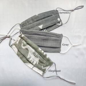 May include: Three face masks with adjustable ear loops are displayed. One is gray with white arrows and geometric shapes, another is solid gray, and the third is green and white with an elephant pattern. The text "Arrows", "Gray", and "Elephant" are visible.