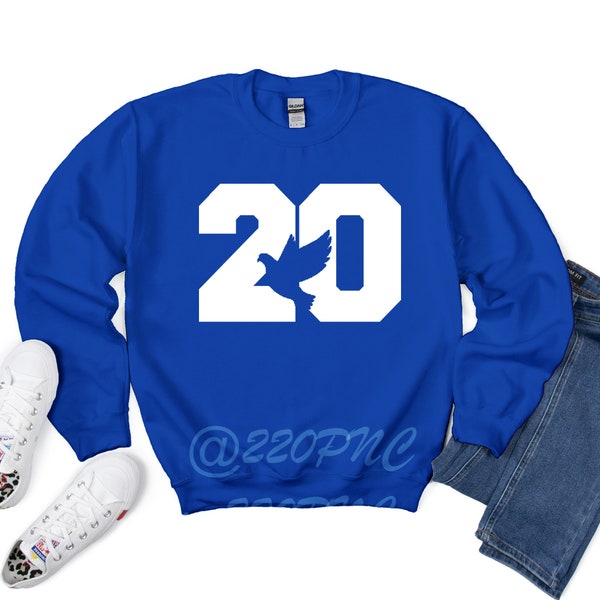 Op de afbeelding: Een koninklijk blauw sweatshirt met witte letters die "20" zeggen met een witte duif die door de nul vliegt. Het sweatshirt heeft de tekst "@22opnc" onderaan.