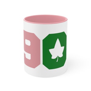 Puede incluir: Taza de cerámica blanca con borde rosa. La taza tiene un "9" rosa y un diseño de hoja verde.
