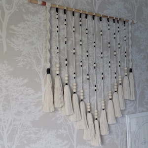 Peut inclure: Une suspension murale en macramé blanche avec des perles noires et blanches et des glands. La suspension est faite de cordon de coton et est fixée à une baguette en bois.