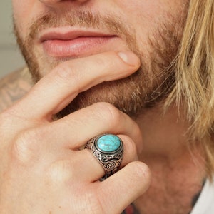 Könnte beinhalten: Silberfarbener Ring mit einem ovalen türkisfarbenen Stein. Der Ring hat ein verziertes, wirbelndes Design um den Stein herum. Die Person trägt ein weißes Tanktop.