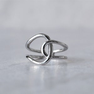 Puede incluir: Un anillo de plata con un dise&ntilde;o &uacute;nico e entrelazado. El anillo est&aacute; hecho de una sola pieza de metal que se dobla y tuerce para crear una forma de nudo.