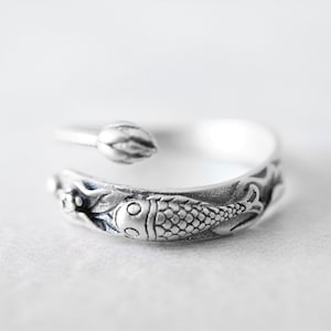 Può includere: Un anello argentato con un design aperto. L'anello presenta un motivo dettagliato di pesce e piante acquatiche. Un piccolo elemento a forma di bocciolo si trova all'estremità opposta del pesce. L'anello è probabilmente in argento sterling.