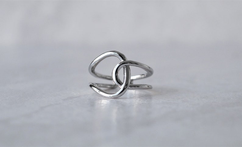 Puede incluir: Un anillo de plata con un dise&ntilde;o &uacute;nico e entrelazado. El anillo est&aacute; hecho de una sola pieza de metal que se dobla y tuerce para crear una forma de nudo.