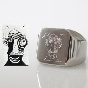 Puede incluir: Un anillo de sello de plata con una cara cuadrada que presenta una ilustración grabada en blanco y negro de un personaje de dibujos animados con ojos grandes.