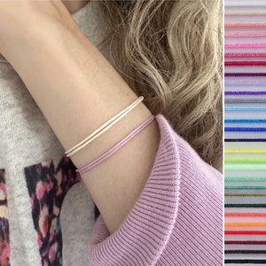 May include: Two minimalist bracelets. One is a light cream colour, and the other is a soft lavender. The bracelets are thin and appear to be made of cord. A colour chart of various cord colours is visible in the background.