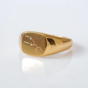 Könnte beinhalten: Ein goldfarbener Siegelring mit polierter Oberfläche. Der Ring hat eine rechteckige Oberfläche mit einem Sternbild-Design. Das Sternbild besteht aus kleinen, weißen Punkten. Ein klassisches Schmuckstück.