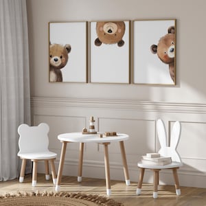 May include: A white wooden table and two chairs in a child's playroom. The table has a round top and four legs. The chairs are shaped like a bear and a bunny. There are three framed prints on the wall behind the table, each featuring a cartoon bear.