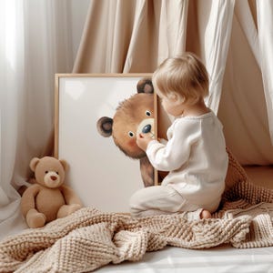 May include: A child in a white onesie is looking at a framed illustration of a brown bear with a pink nose. The bear is peeking out from behind the frame. A brown teddy bear is sitting on a beige knitted blanket.